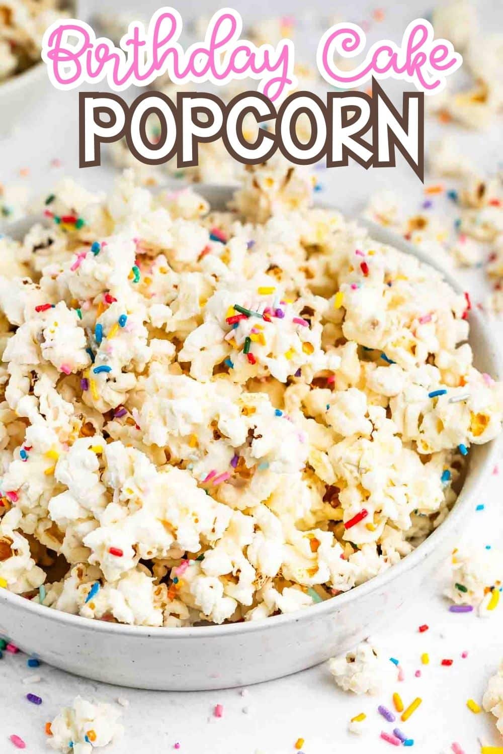A bowl of popcorn covered in white coating and colorful sprinkles sits on a white surface. Text above the bowl reads Birthday Cake Popcorn in pink and brown letters.