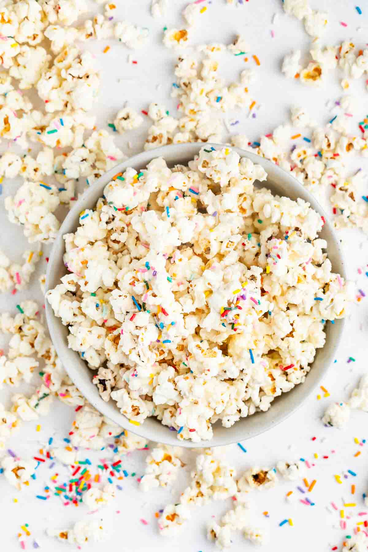 A bowl of popcorn topped with colorful rainbow sprinkles, with more popcorn and sprinkles scattered on a white surface around the bowl.