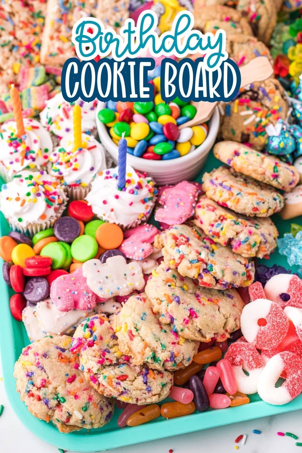 A colorful birthday cookie board featuring sprinkle cookies, frosted cupcakes with candles, a bowl of candy-coated chocolates, and assorted gummy candies on a turquoise tray. The text “Birthday Cookie Board” is at the top.