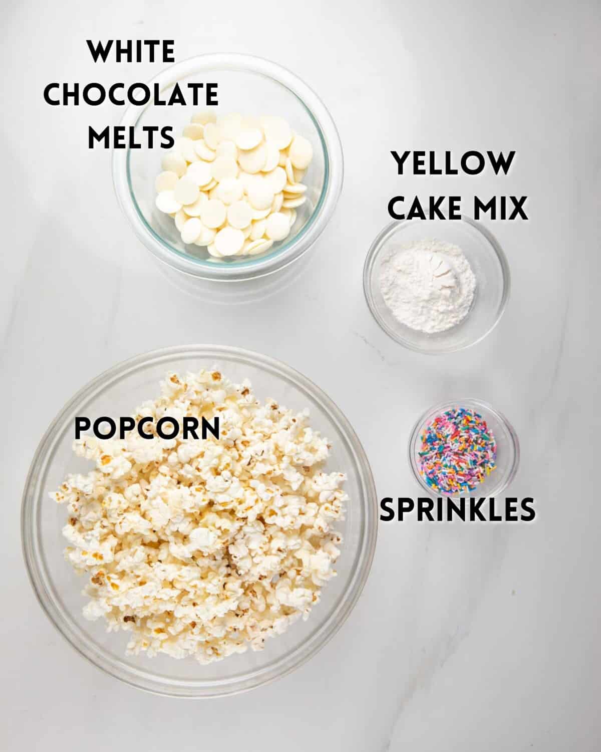 Four bowls on a white surface contain white chocolate melts, yellow cake mix, popcorn, and colorful sprinkles, each labeled with bold text.