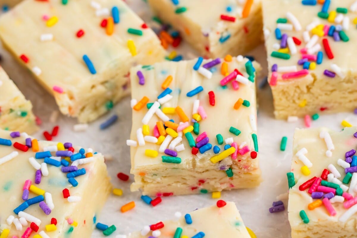 Close-up of creamy white fudge squares topped with colorful rainbow sprinkles, arranged on a white parchment surface. The fudge looks rich and festive, with a smooth texture and fun, vibrant colors.