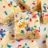 Close-up of creamy white fudge squares topped with colorful rainbow sprinkles, arranged on a white parchment surface. The fudge looks rich and festive, with a smooth texture and fun, vibrant colors.