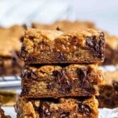 Three gooey caramel and chocolate chip cookie bars are stacked on top of each other, with melted chocolate and caramel oozing out. Pieces of caramel candy are scattered around on a white surface.