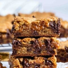 Three gooey caramel and chocolate chip cookie bars are stacked on top of each other, with melted chocolate and caramel oozing out. Pieces of caramel candy are scattered around on a white surface.