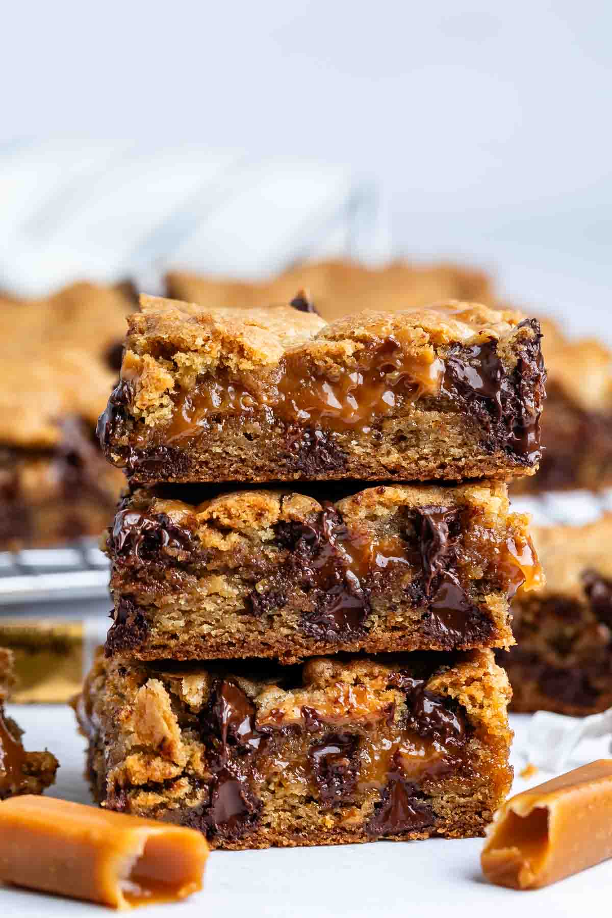 Three gooey caramel and chocolate chip cookie bars are stacked on top of each other, with melted chocolate and caramel oozing out. Pieces of caramel candy are scattered around on a white surface.