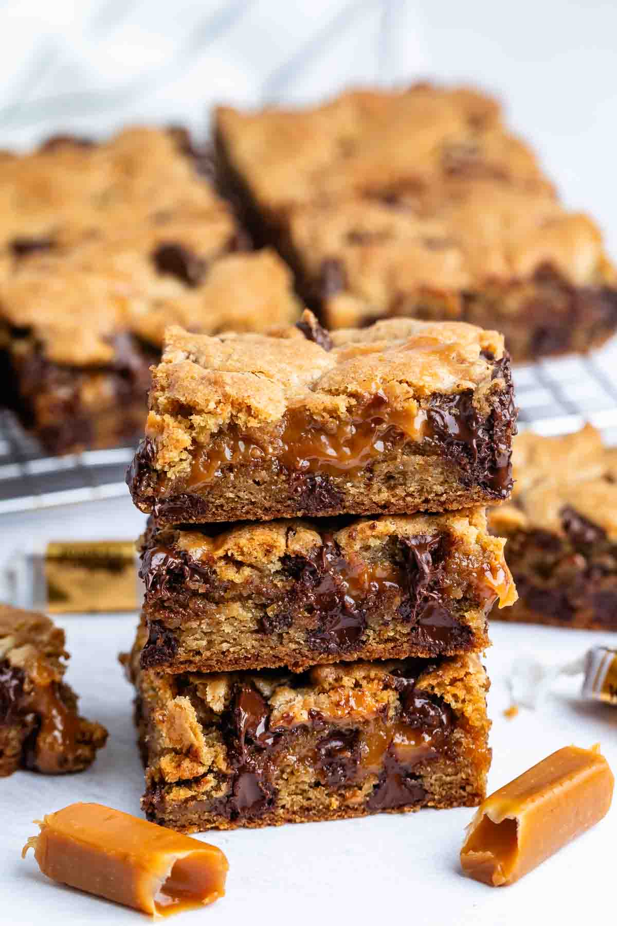 Three caramel-filled chocolate chip cookie bars are stacked in the foreground, showing gooey caramel and melted chocolate. More bars and unwrapped caramel candies are scattered around on a white surface.