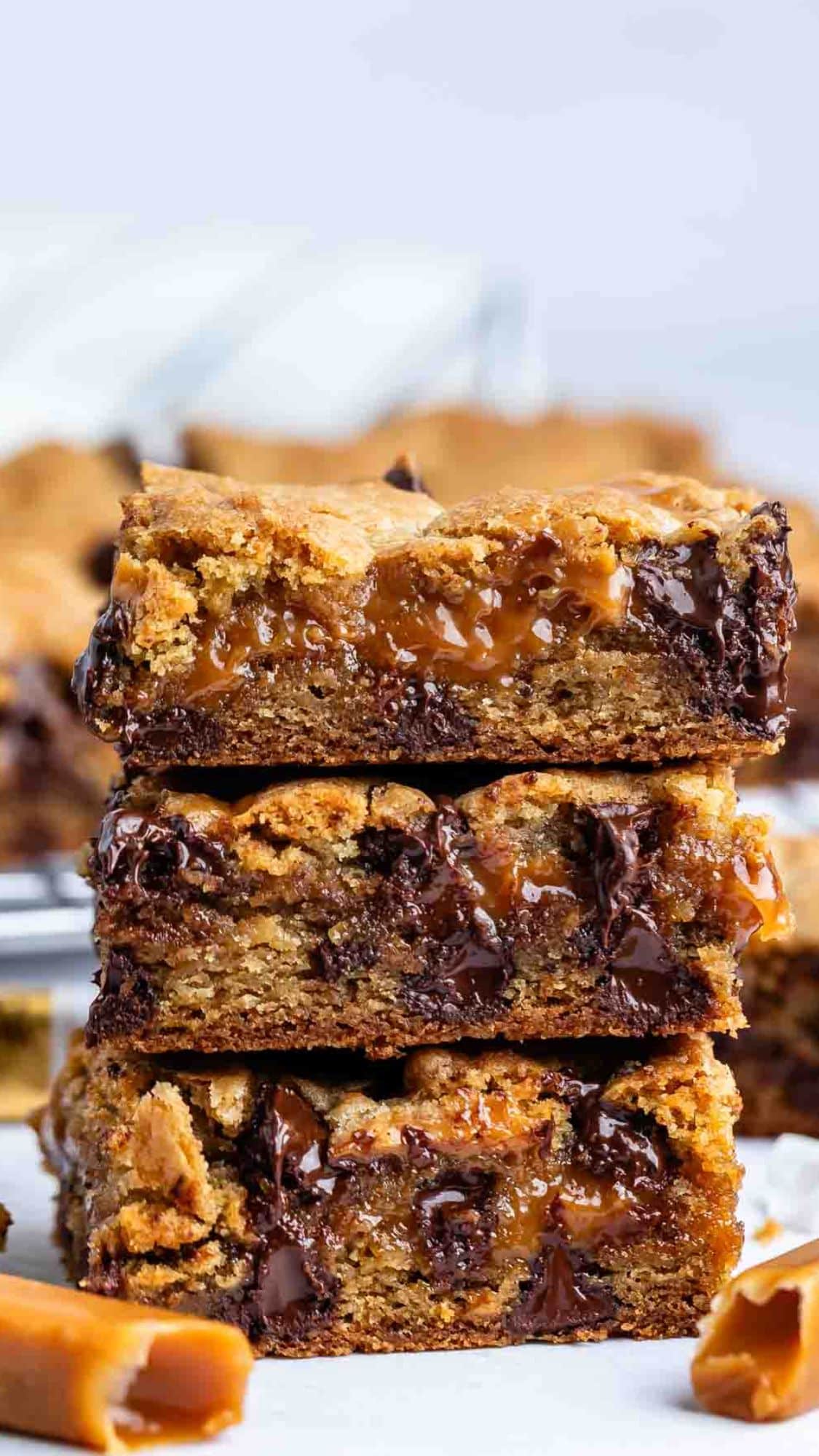 A stack of three gooey caramel and chocolate chip cookie bars, with melted caramel and chocolate oozing from the centers, sits in focus. Pieces of caramel candy are visible in the foreground.