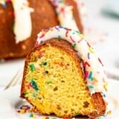 A slice of bundt cake with colorful sprinkles baked inside, topped with white icing and more sprinkles, sits on a white plate with scattered sprinkles around it.