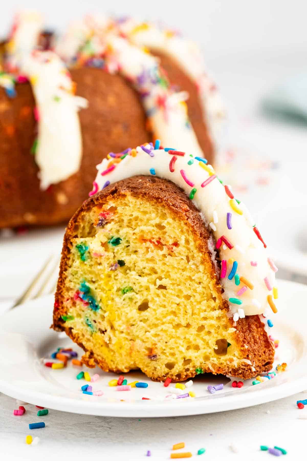 A slice of bundt cake with colorful sprinkles baked inside, topped with white icing and more sprinkles, sits on a white plate with scattered sprinkles around it.