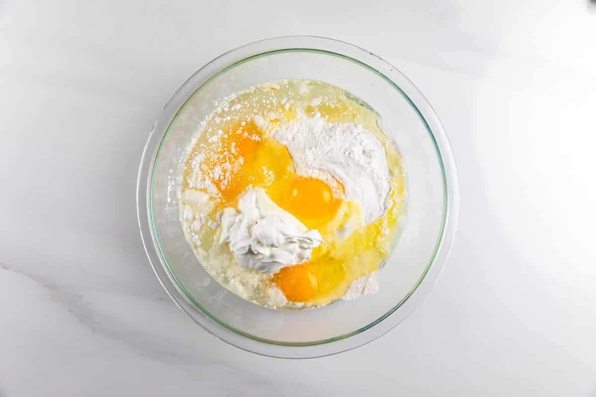 A glass mixing bowl with flour, three cracked eggs, a dollop of yogurt or sour cream, and water on a white surface, ready to be mixed.