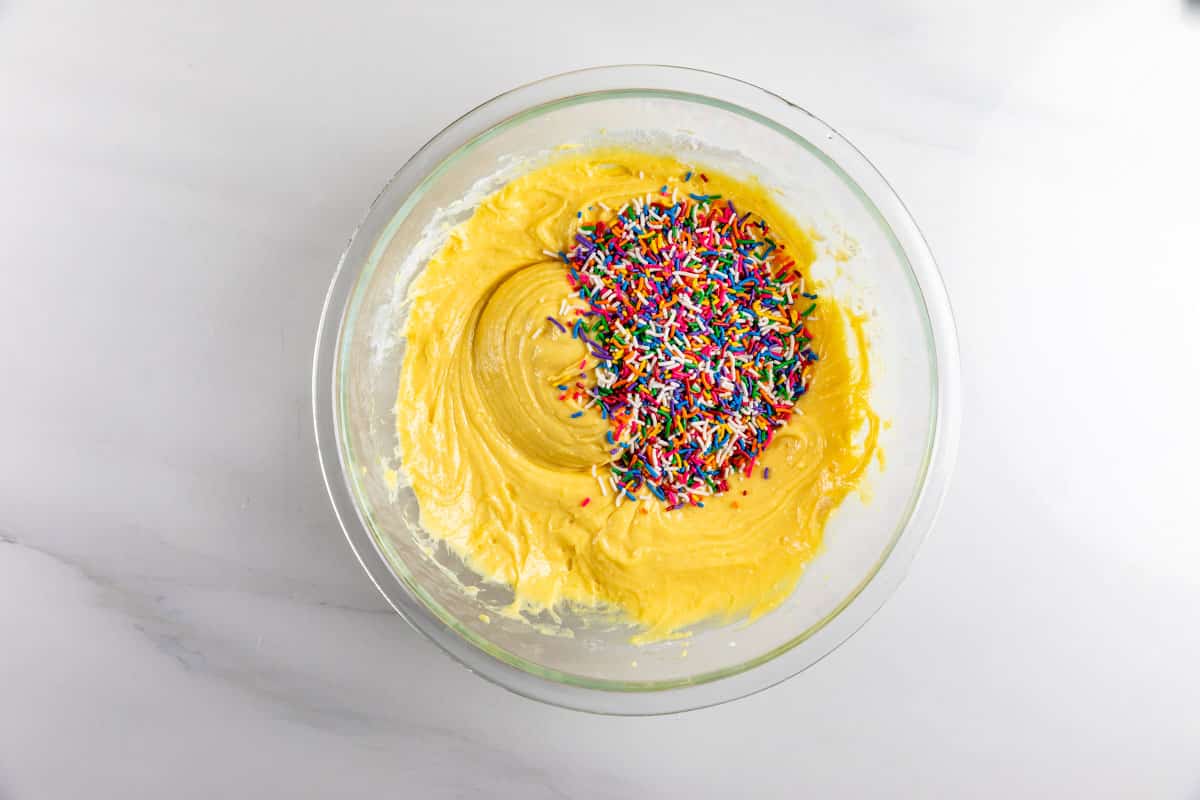 A glass bowl filled with yellow cake batter sits on a white surface. Colorful rainbow sprinkles are piled on top of half of the batter, not yet mixed in.