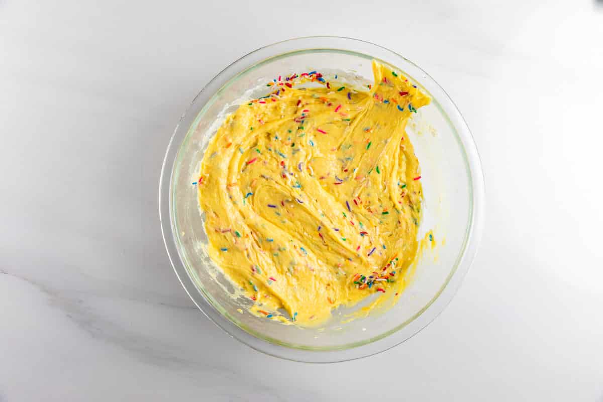 A glass bowl filled with yellow cake batter mixed with colorful rainbow sprinkles, sitting on a white marble surface.