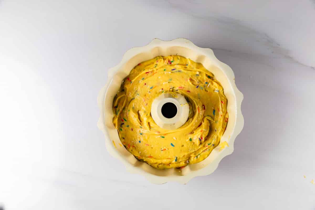 A bundt cake pan filled with yellow cake batter mixed with colorful sprinkles, sitting on a white marble surface.