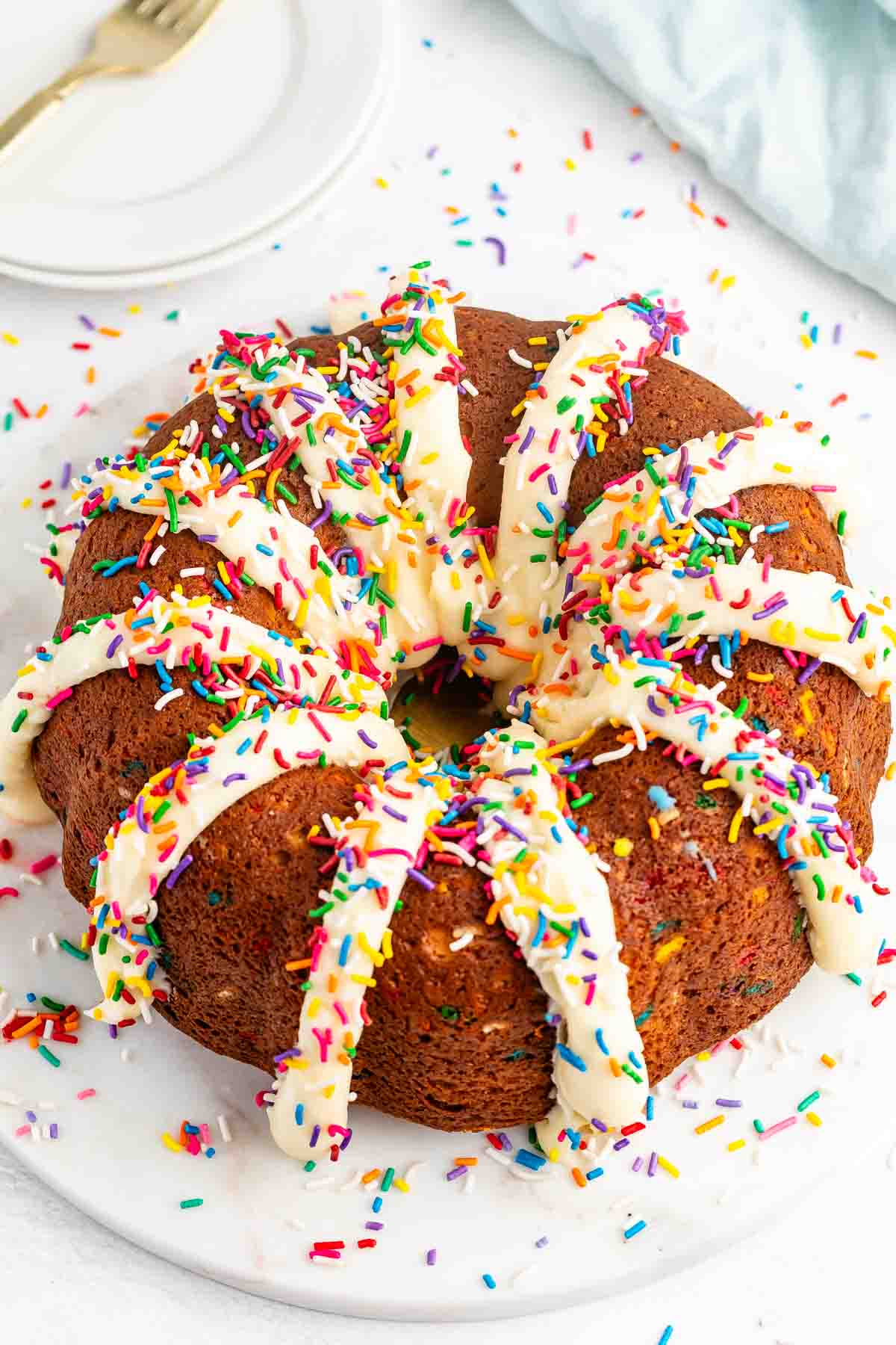 A round bundt cake topped with white icing and colorful rainbow sprinkles sits on a white marble surface, with more sprinkles scattered around. A plate and fork are visible in the background.