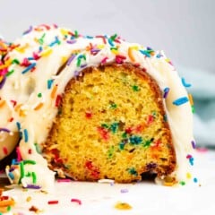 A close-up of a sliced bundt cake with colorful sprinkles mixed inside, topped with white icing and more rainbow sprinkles on top and around the cake.