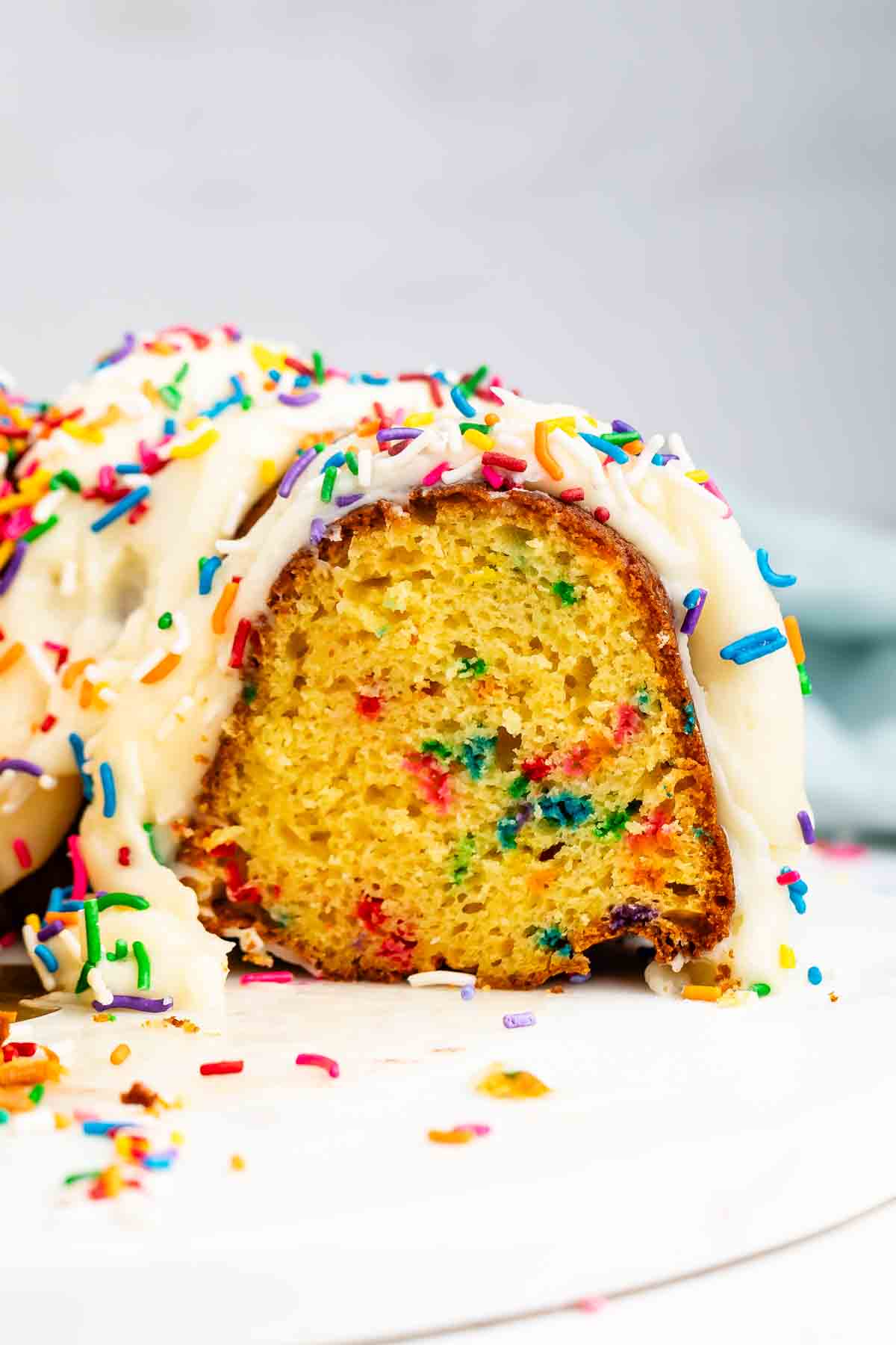 A close-up of a sliced bundt cake with colorful sprinkles mixed inside, topped with white icing and more rainbow sprinkles on top and around the cake.