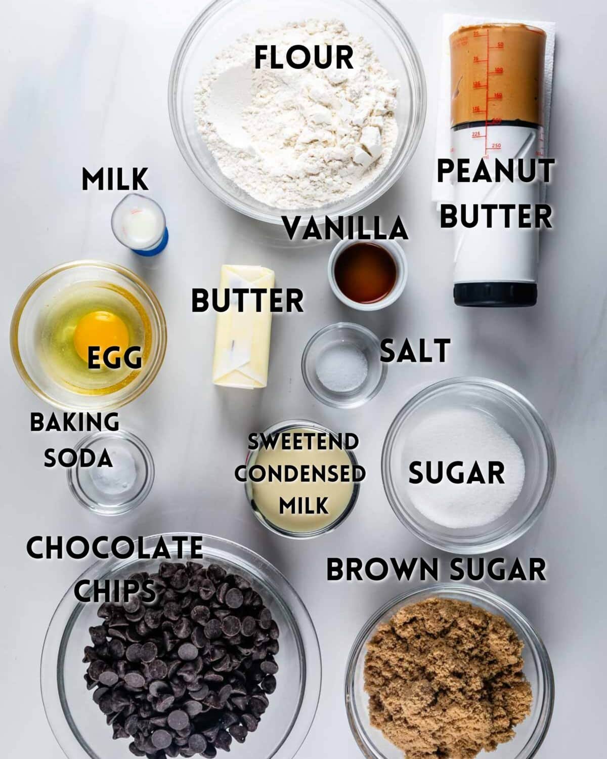 Overhead view of baking ingredients in bowls on a white surface, including flour, milk, egg, butter, baking soda, sweetened condensed milk, peanut butter, vanilla, salt, sugar, brown sugar, and chocolate chips. Labels identify each ingredient.