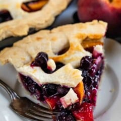 A slice of fruit pie with a lattice crust, filled with blueberries and peach slices, sits on a white plate with a fork. A whole peach and the remaining pie are in the background.