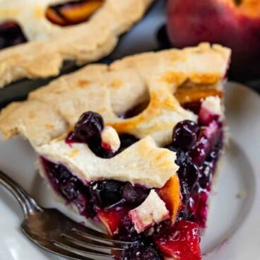 A slice of fruit pie with a lattice crust, filled with blueberries and peach slices, sits on a white plate with a fork. A whole peach and the remaining pie are in the background.