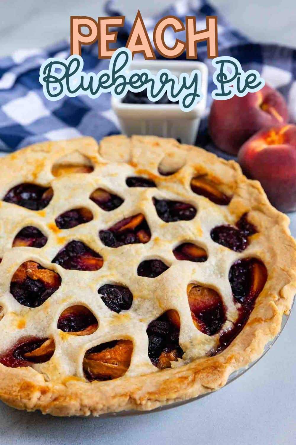 A peach and blueberry pie with a golden crust featuring round cutouts, set on a table with a blue checkered cloth and two whole peaches in the background. Text overlay reads Peach Blueberry Pie.