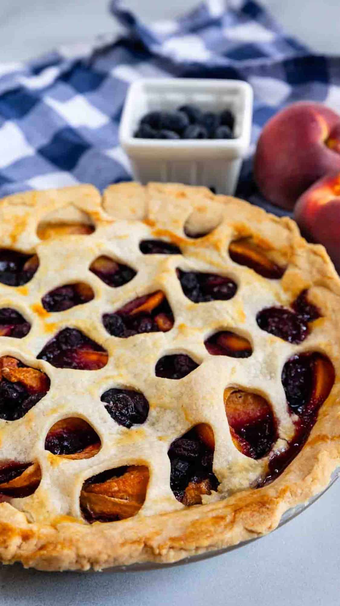 A freshly baked peach and blueberry pie with a lattice crust sits on a table. Behind it are a blue-and-white checkered cloth, a bowl of blueberries, and two whole peaches.