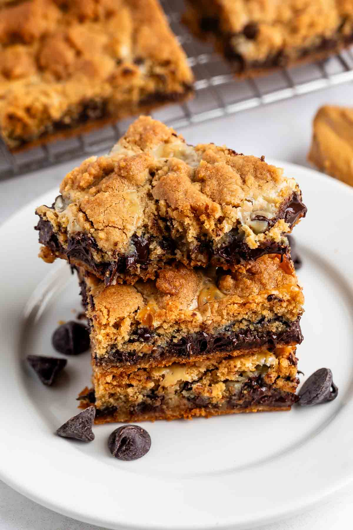 Three chocolate chip cookie bars stacked on a white plate, with chocolate chips scattered around. The bars have a gooey center and golden-brown, crumbly tops. More bars are visible on a cooling rack in the background.