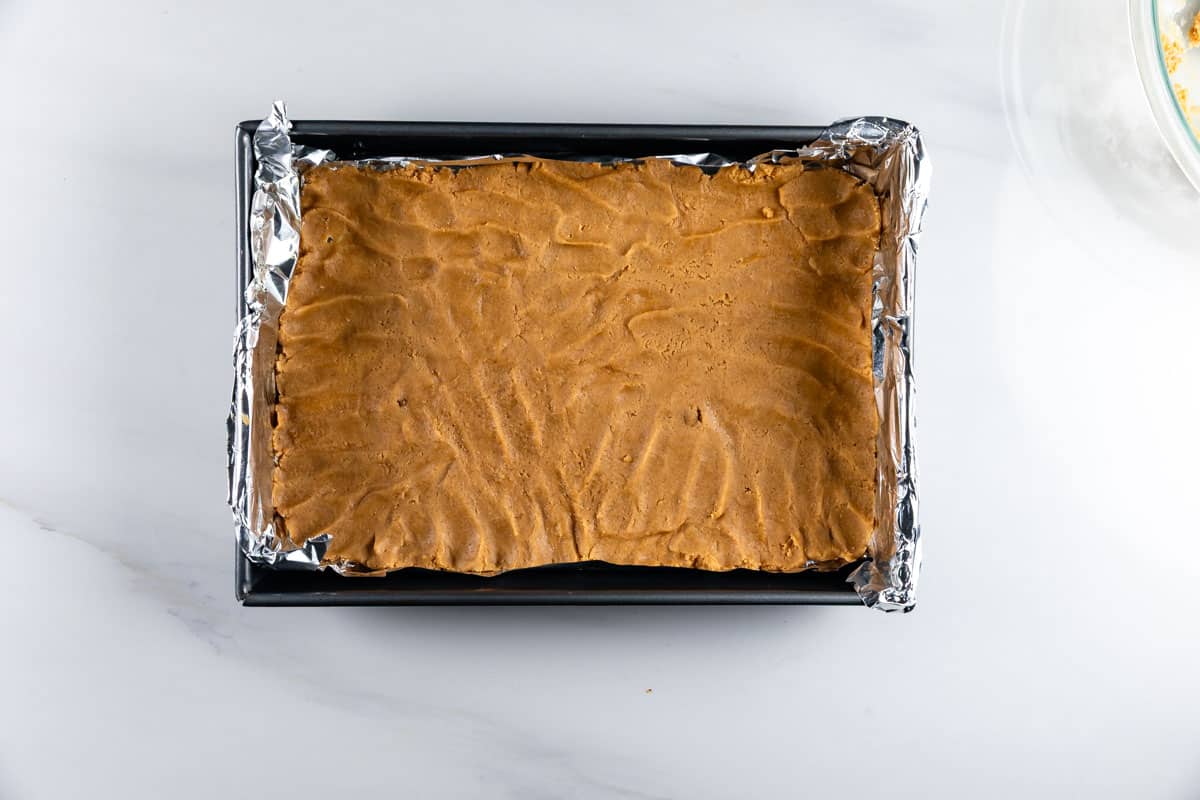 A rectangular baking pan lined with aluminum foil contains an unbaked, evenly pressed layer of light brown dough on a white surface.