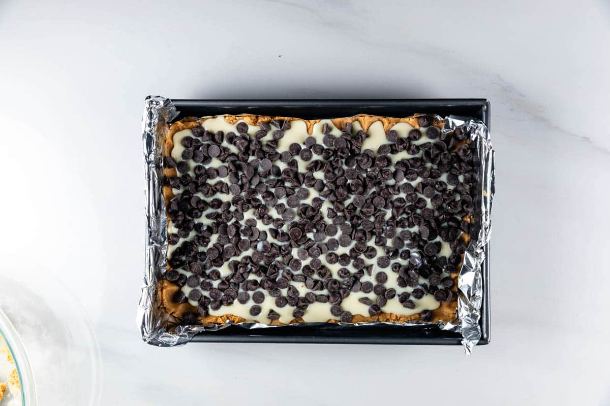 A rectangular baking pan lined with foil contains a dessert topped with a layer of chocolate chips and a white creamy layer underneath, set on a white marble surface.