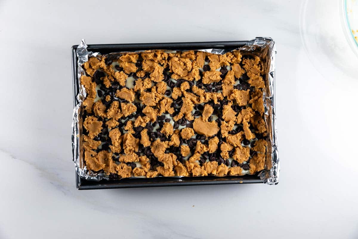 A rectangular baking pan lined with foil contains an unbaked layered dessert topped with chunks of cookie dough and chocolate chips on a white surface.
