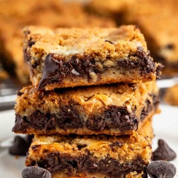 A stack of three gooey chocolate chip cookie bars on a white plate, with melted chocolate visible and a few chocolate chips scattered around. The background shows more cookie bars slightly out of focus.