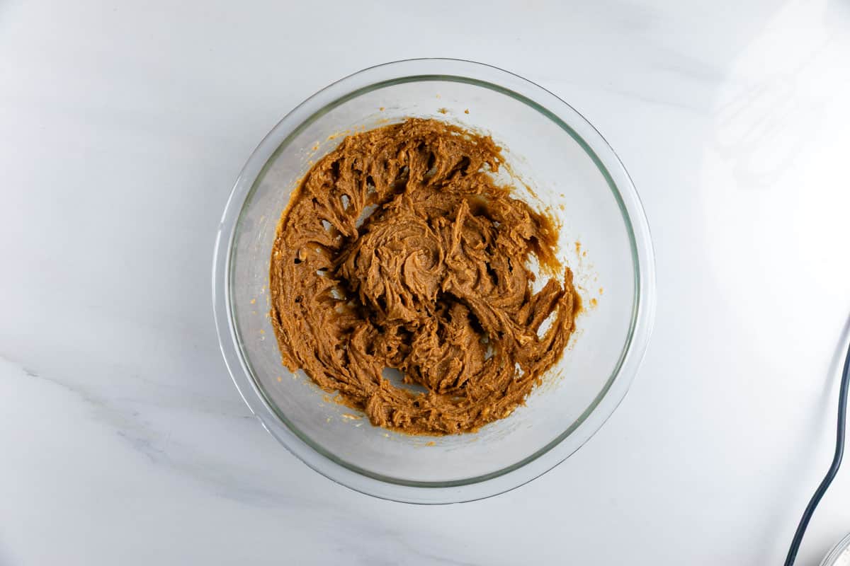 A glass mixing bowl on a white countertop filled with a thick, brown cookie dough mixture, partially mixed.