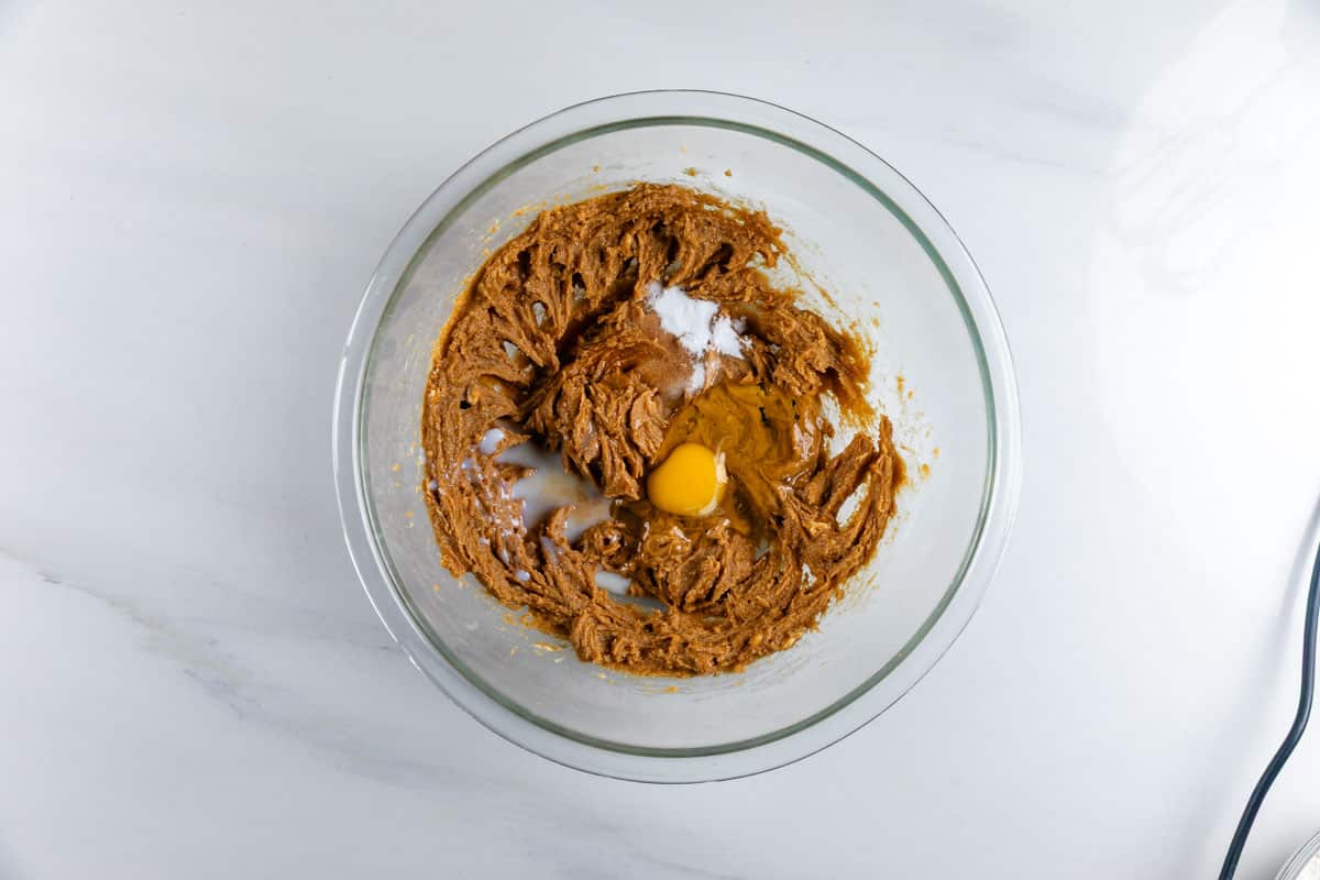 A clear glass mixing bowl contains brown cookie dough, an egg, and a bit of white sugar on top, sitting on a white marble countertop.