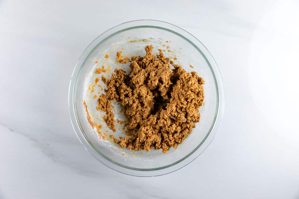 A clear glass mixing bowl filled with light brown cookie dough sits on a white marble surface.