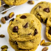 A stack of chocolate chip cookies with visible pistachios sits on a white surface, surrounded by scattered pistachios and more cookies cooling on a wire rack in the background.