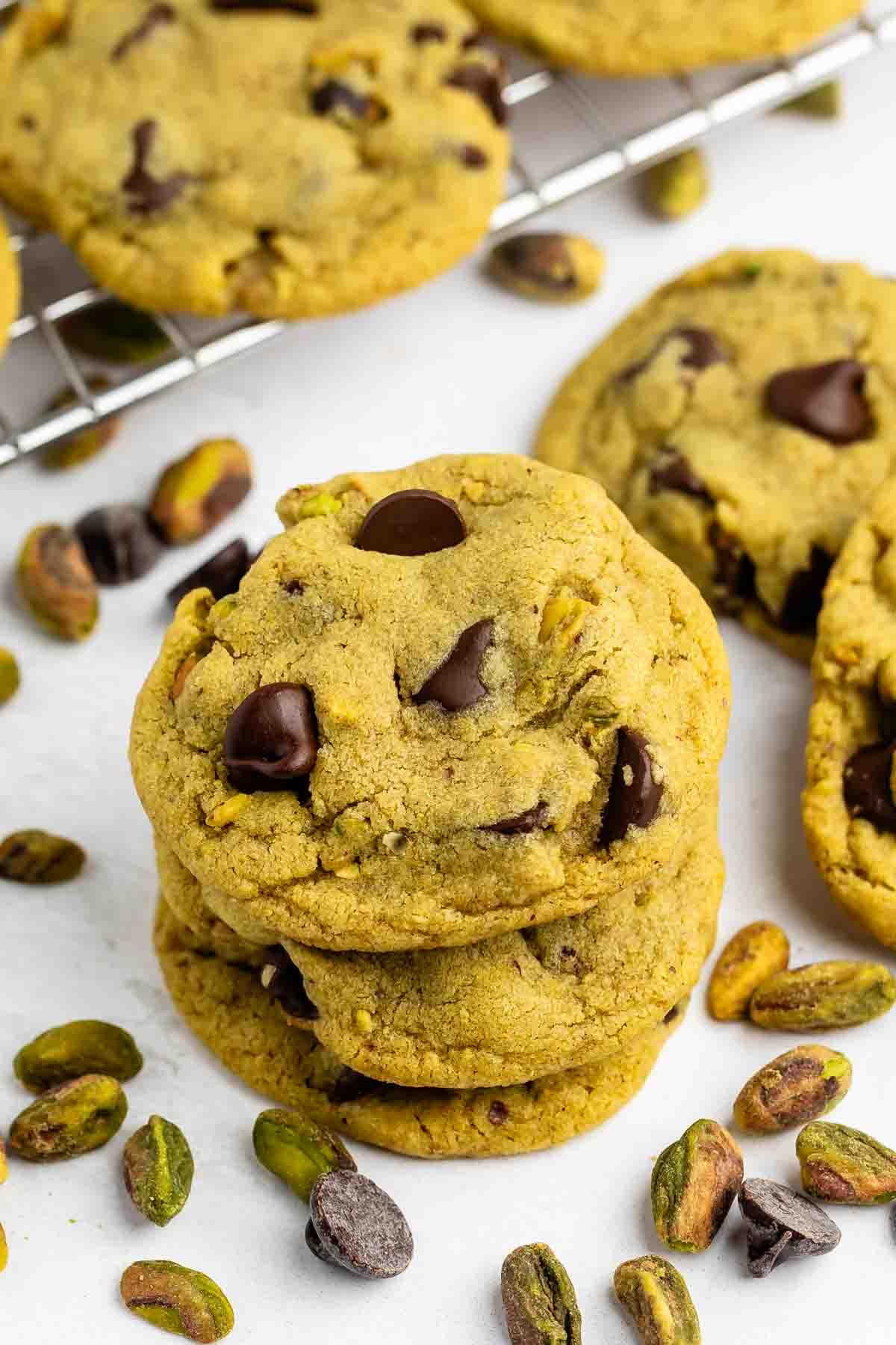 A stack of chocolate chip cookies with visible pistachios sits on a white surface, surrounded by scattered pistachios and more cookies cooling on a wire rack in the background.