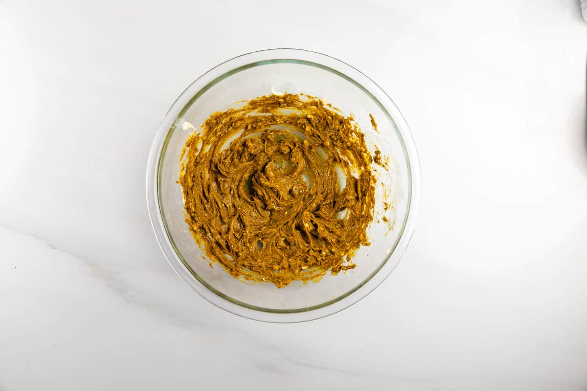 A glass bowl containing thick, brownish cookie dough mixture sits on a white marble surface.
