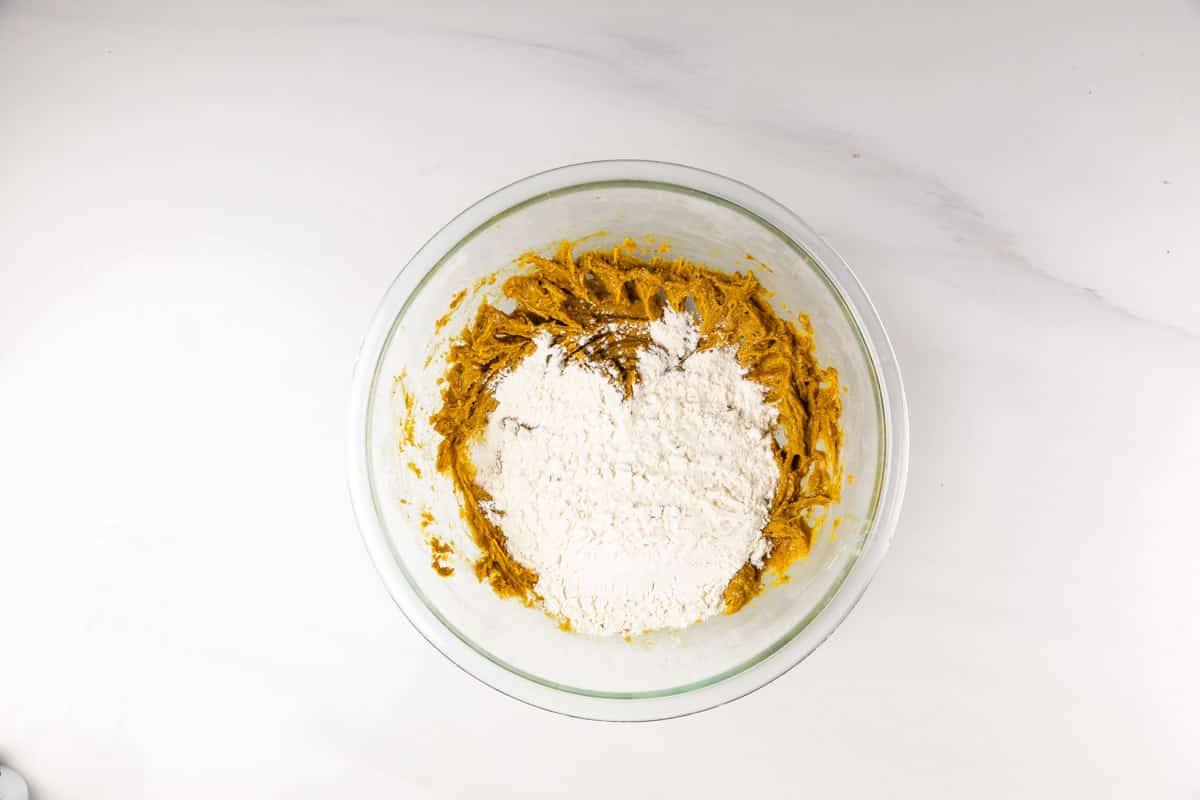 A glass mixing bowl on a white surface contains partially mixed cookie dough with a mound of flour on top.