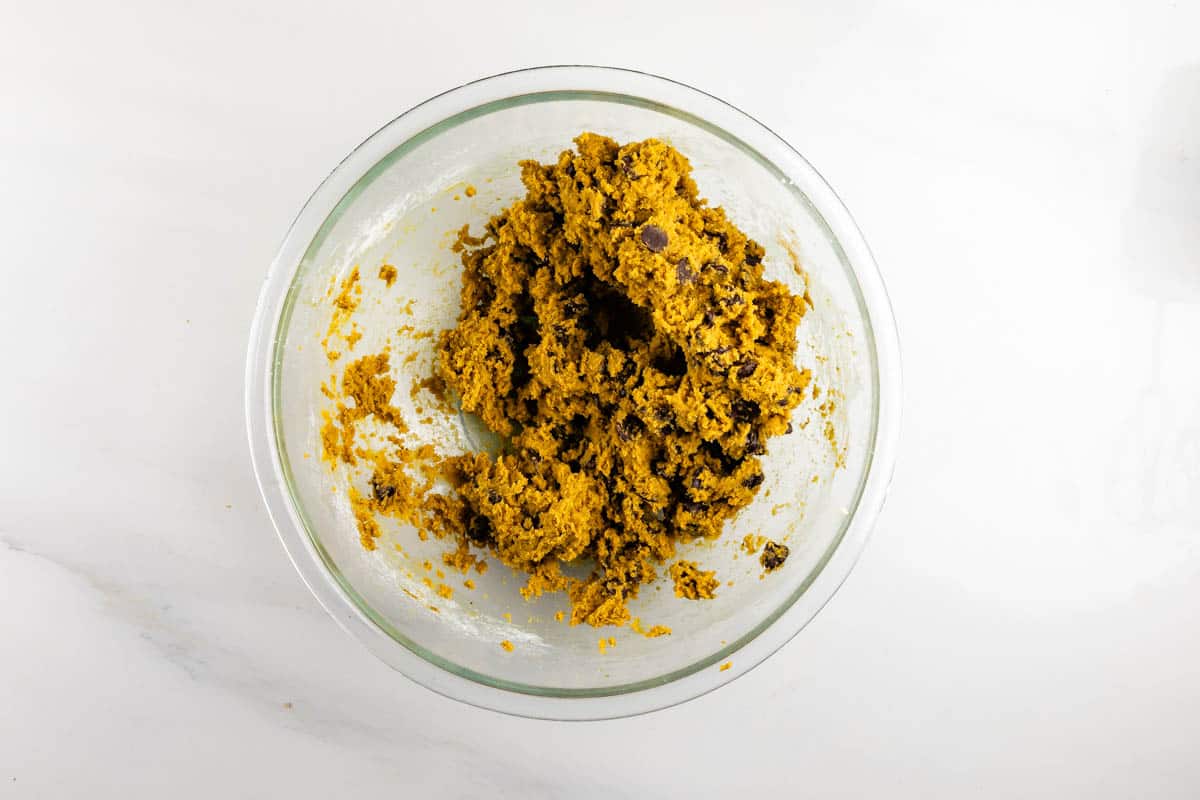 A glass bowl containing orange-brown cookie dough with chocolate chips, set on a white countertop.