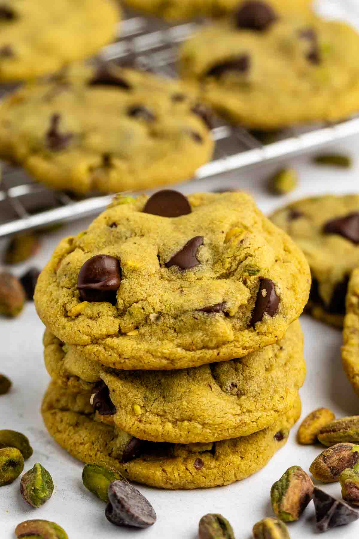 A stack of three chocolate chip cookies with visible chunks of chocolate and pistachios, surrounded by more cookies and scattered pistachios on a white surface. A cooling rack with more cookies is in the background.