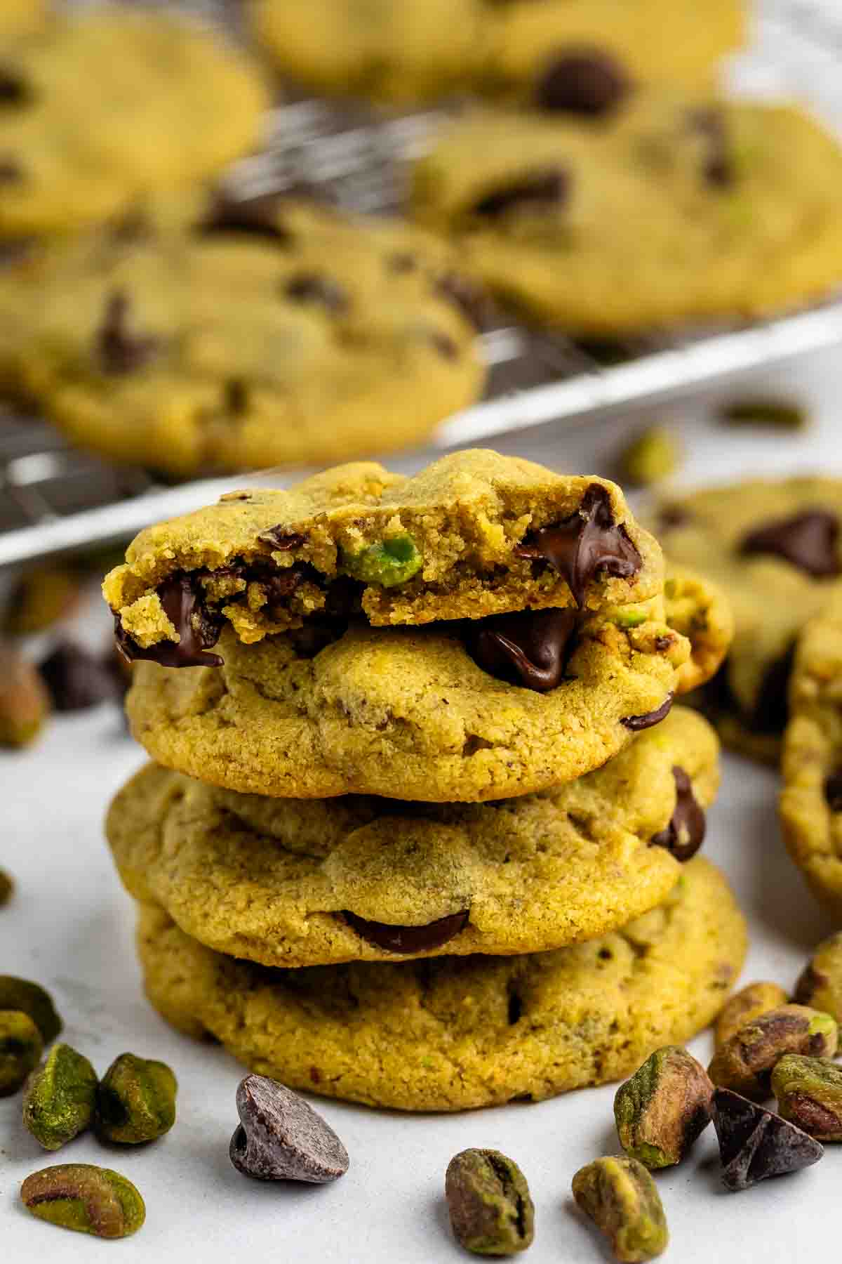 A stack of three chocolate chip cookies with green pistachio pieces, topped with a cookie that has a bite taken out. More cookies and scattered pistachios are visible in the background.