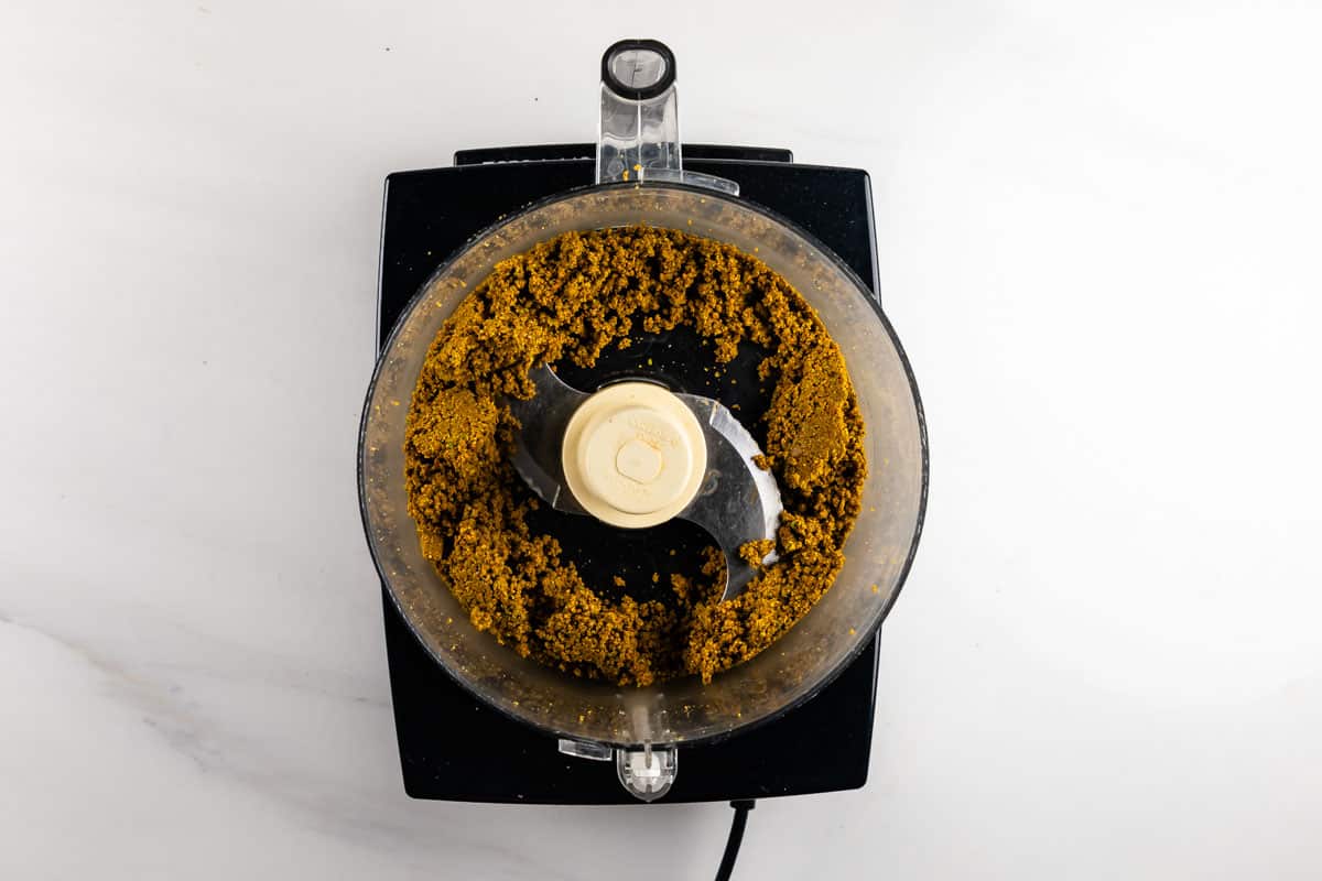 Overhead view of a food processor containing a coarse, brownish mixture, sitting on a white countertop. The mixture is partially spread around the sides of the bowl.