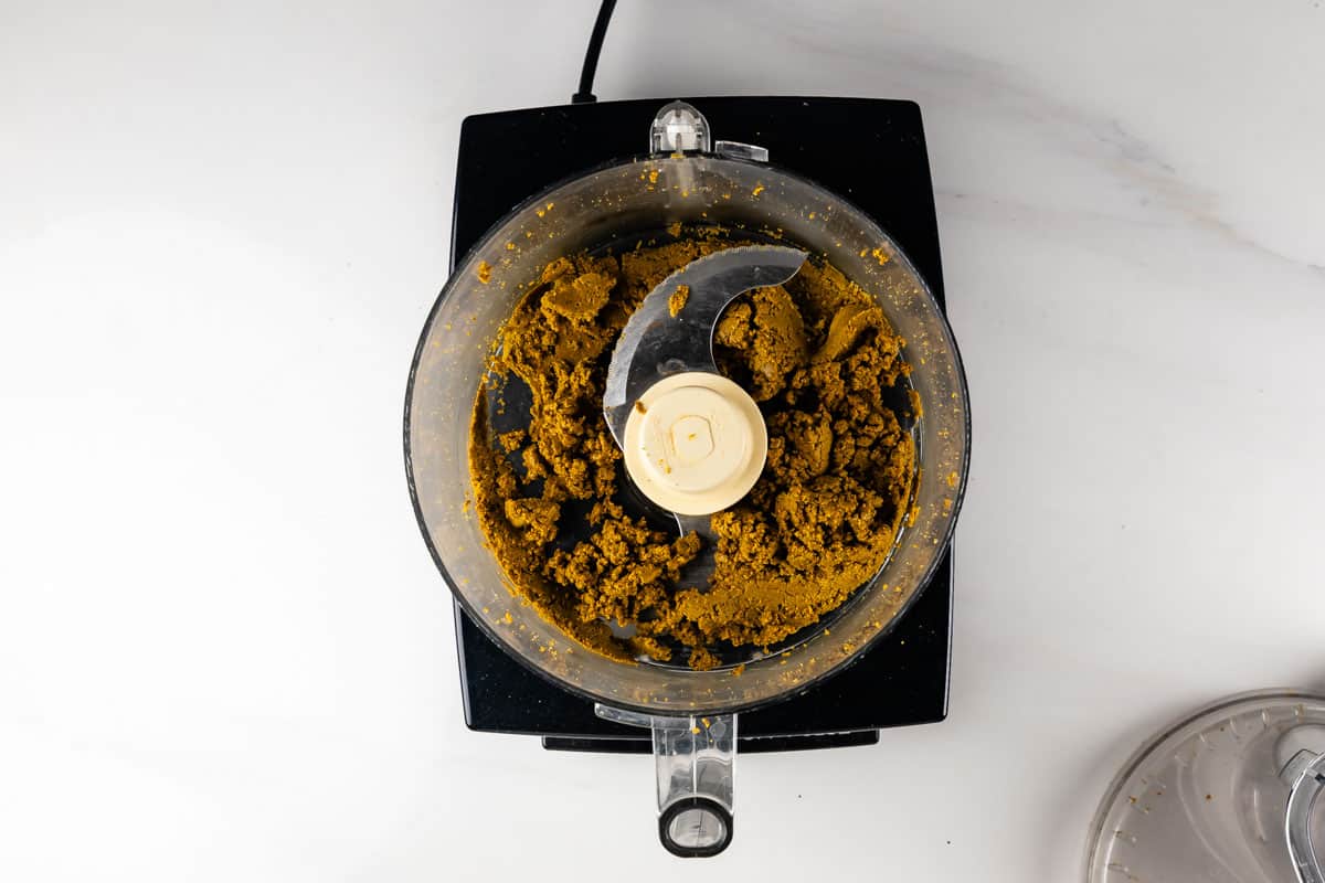 Overhead view of a food processor containing a thick, brownish mixture with a blade in the center, sitting on a white countertop.