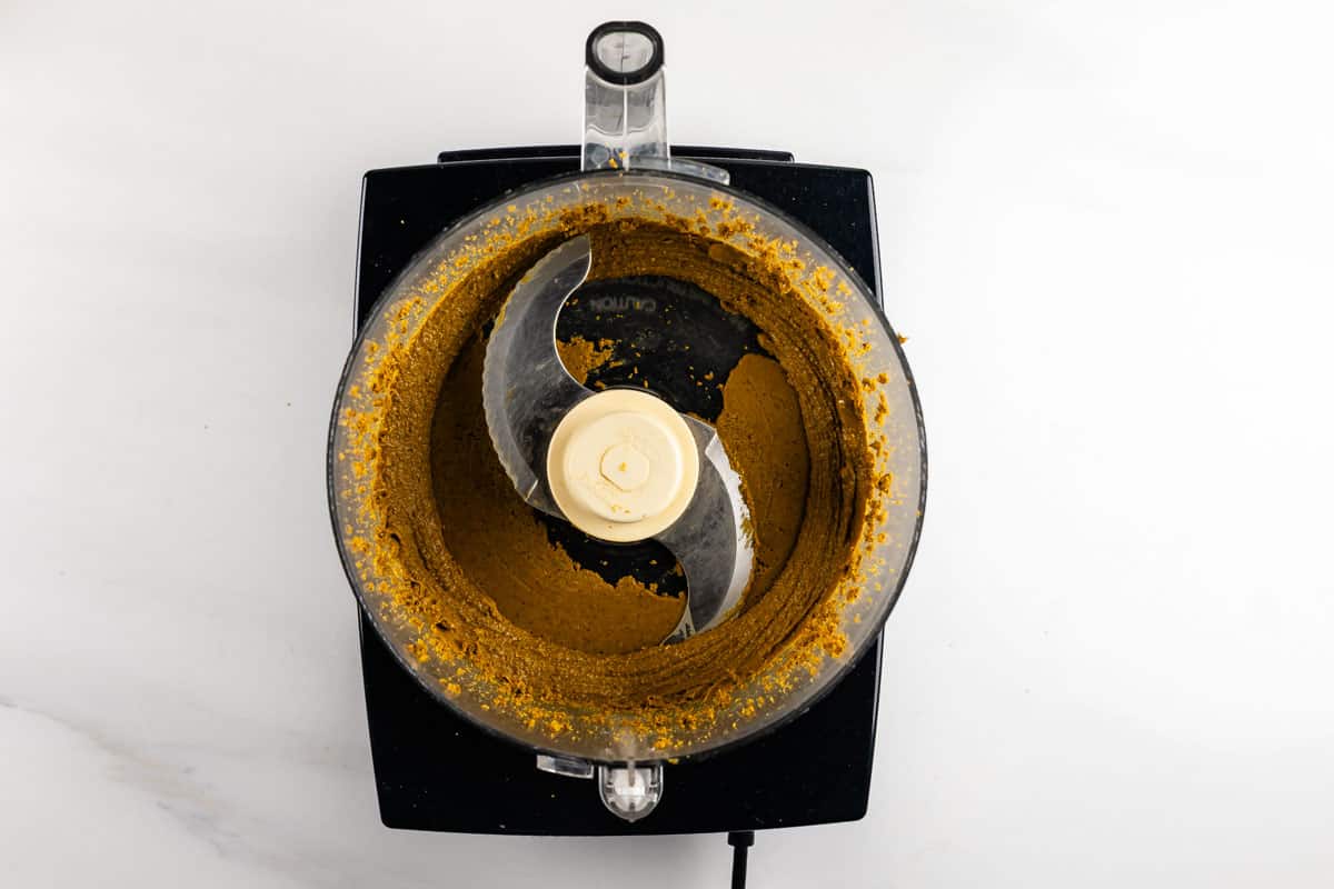 Overhead view of a food processor containing a finely ground, yellowish-brown mixture spread around the bowl, with the central blade visible. The processor is on a white surface.