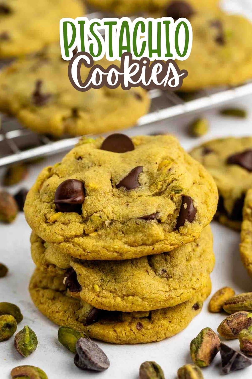 A stack of three pistachio cookies with chocolate chips sits on a white surface, surrounded by scattered pistachios. More cookies and a cooling rack are in the background. Text above reads “Pistachio Cookies.”.