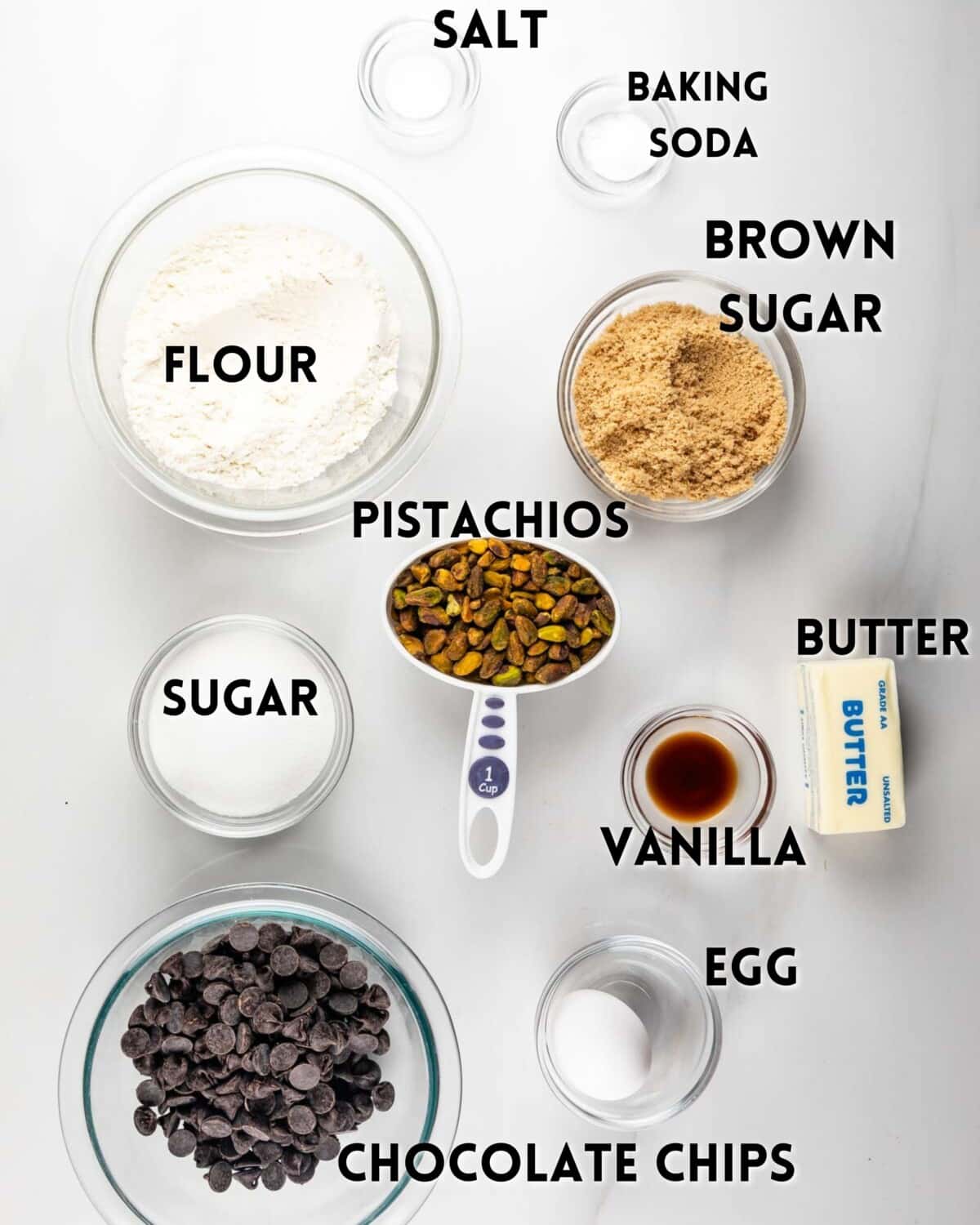 Top-down view of baking ingredients in bowls on a white surface, labeled flour, brown sugar, sugar, pistachios, chocolate chips, butter, vanilla, egg, salt, and baking soda.