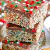 Three stacked crispy cereal bars with a chocolate topping, decorated with red, green, and white holiday sprinkles. A spoonful of peanut butter sits nearby, suggesting a key ingredient.
