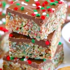 Three stacked crispy cereal bars with a chocolate topping, decorated with red, green, and white holiday sprinkles. A spoonful of peanut butter sits nearby, suggesting a key ingredient.
