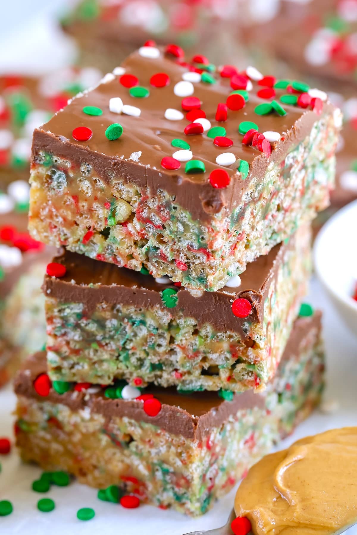 Three stacked crispy cereal bars with a chocolate topping, decorated with red, green, and white holiday sprinkles. A spoonful of peanut butter sits nearby, suggesting a key ingredient.
