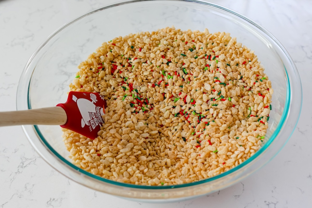 A glass bowl filled with Rice Krispies cereal mixed with colorful sprinkles, being stirred with a red spatula on a white marble countertop.