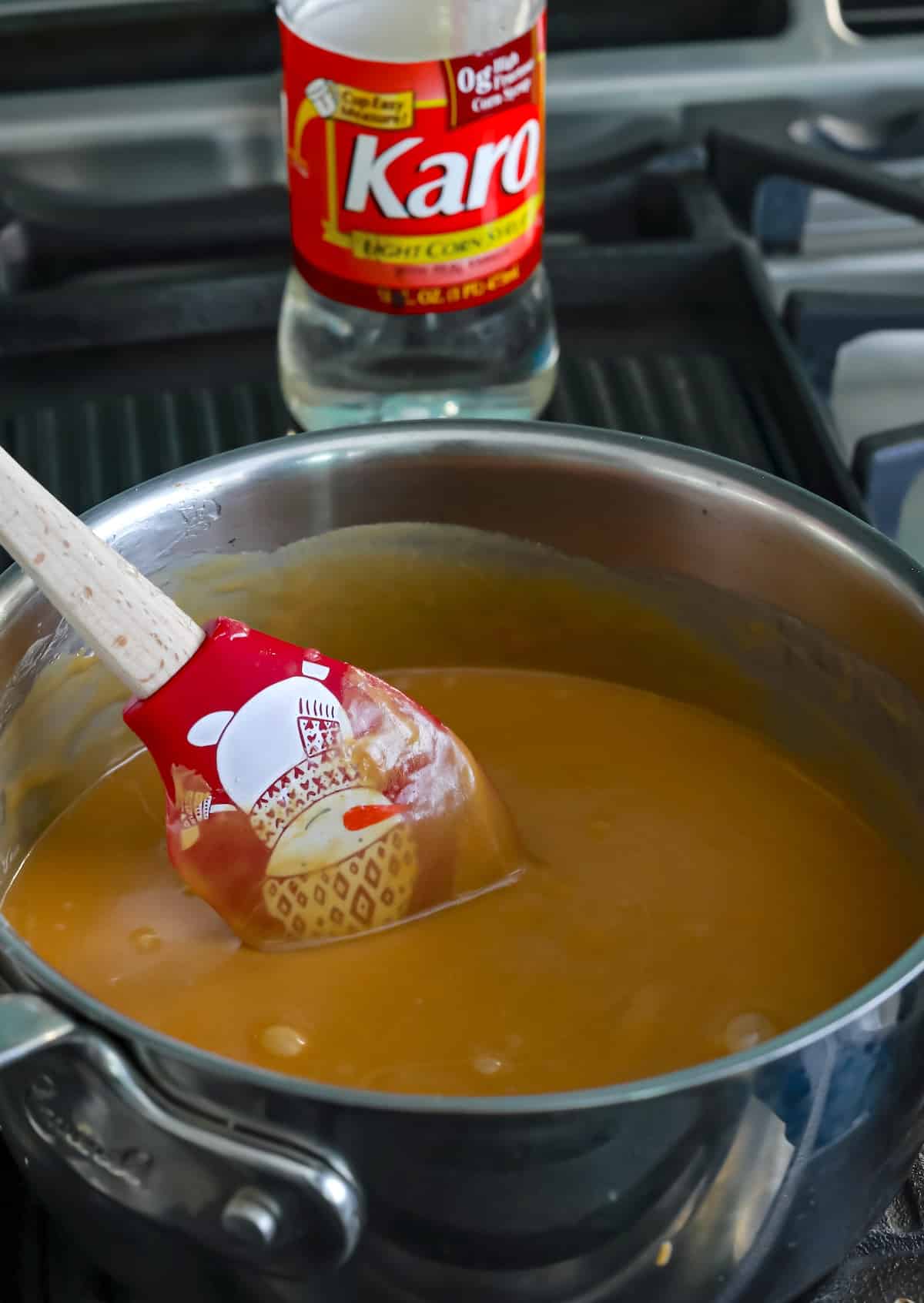 A saucepan filled with caramel mixture being stirred by a red and white spatula on a stove, with a bottle of Karo corn syrup in the background.
